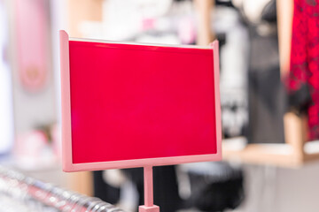 A close-up of a pink clothing rack with a blank red sign. The background features blurred clothing items in a retail store setting.