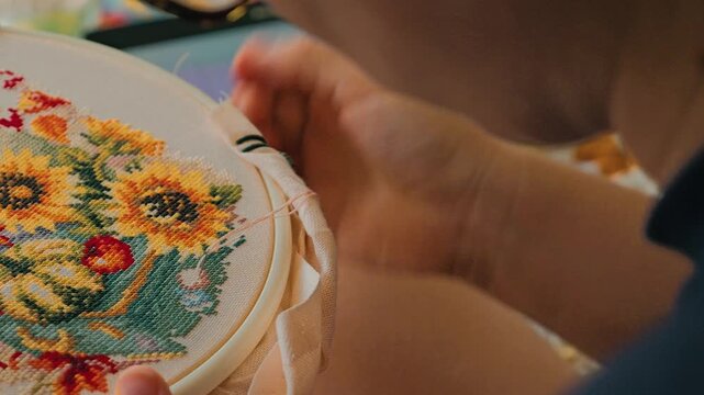 A close-up of hands working on a vibrant embroidery project featuring sunflowers and other floral motifs, with a digital tablet visible in the background. Cross-stitch