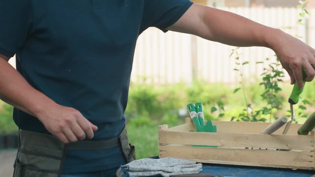 Side view gardener removing tools, dropping on blue drum, dusting hand, apron belt on waist, wooden crate with green handles, gloves visible, yard house fence background