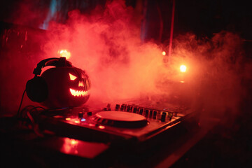 A carved Halloween pumpkin with glowing eyes and headphones sits on a DJ turntable, illuminated by red smoke and lights.