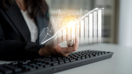 A businesswoman holds a light bulb in one hand while analyzing data on a laptop keyboard. This...