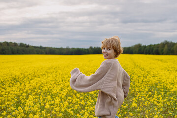 Fototapeta premium Cheerful Caucasian girl, about 10 years old, red hair, wearing beige sweater and jeans, running in yellow flower field, rear view, look at camera, concept childhood, joy, spring, freedom.
