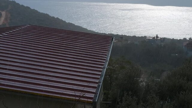 Red roof with sea view under cloudy sky. Raindrops cascading over rusty red metal roofing, streaming along gutters with coastal hills and lush vegetation beneath overcast skies, creating atmospheric