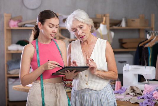 Woman tailor talk with senior female customer and using tablet, enters clients data into tablet for automatic construction of pattern. Designer records clients data in online log..