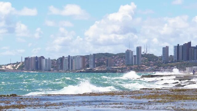 Waves crash on the shore in front of a city skyline on a sunny day