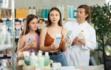 Mother and daughter choose skin cream in a drugstore. Male pharmacist offers to buy skin cream