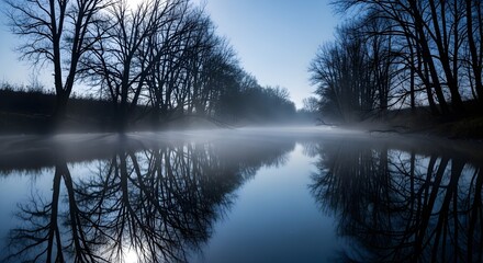Fototapeta premium Misty Winter Morning over a Serene River Lined with Silhouetted Bare Trees and Reflections