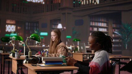 Girl pushing her colleague with physical impairment through the library, studying for a new exam with academic materials from the campus database. African american student in wheelchair. Camera B.
