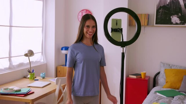 Teenage student girl, 15 years old, recording herself while talking to the camera using a smartphone in her bedroom. She is creating social media content through reels, live streaming and vlogs
