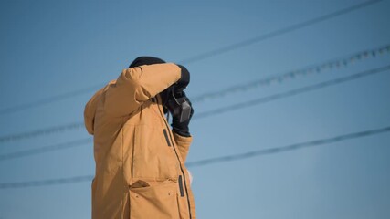 lower view of nature documentarian peering through camera view gloved hands delicately adjusting focus dial then scanning urban winter landscape under bright sunshine clear sky capturing details