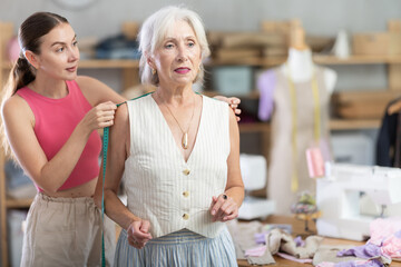 Adult woman seamstress taking body measurements of elderly woman with measuring tape in sewing workshop