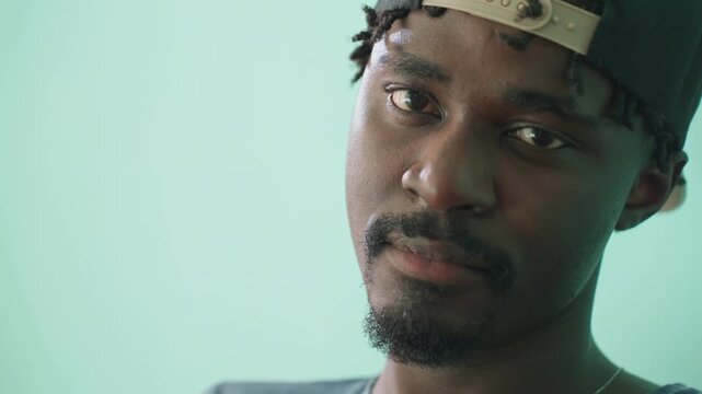 Close up of young man turning head toward camera after staring pensively, lost in thought, wearing cap backwards, showing contemplative expression on face against teal background in soft light