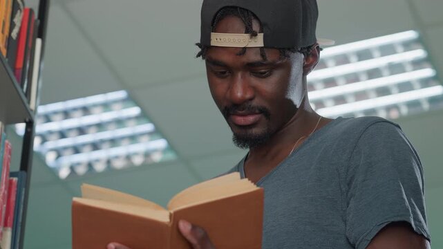 Close up of young man in casual shirt and backwards cap reading hardcover book in library under bright fluorescent lights with focused expression, holding pages carefully