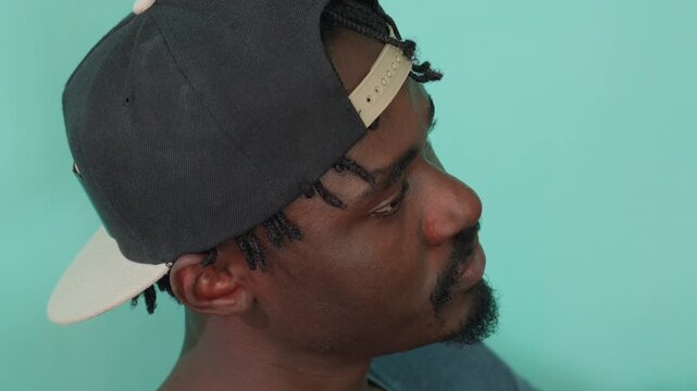 Close up side profile of dark skin man wearing black backward cap looking up thoughtfully with calm expression against mint green background