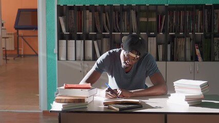 Young man in cap writes notes during study session in library, seated at desk between tall stacks of books, focused under sunlit window, shelves behind, organized materials spread for research