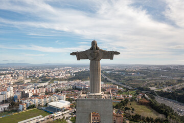 Aerial drone shot of iconic Christ the King statue in Almada, Portugal with arms outstretched overseeing the cityscape under bright sky
