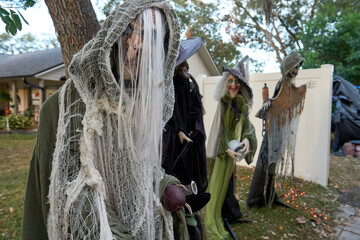 Hooded ghoul stands in the foreground while several witches gather behind a white fence near a house. Outdoor Halloween yard display with orange lights and spooky props.