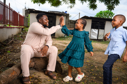 A father sits on a log smiling as he gives a joyful high-five to his daughter, while another child approaches happily from the side. The image captures a lively family bonding moment outdoors.
