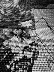 A close-up of a detailed grayscale cross-stitch project, showcasing the intricate stitches and the needle and thread working on the monochromatic pattern.