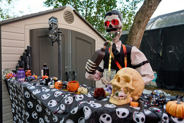 Animated skeleton with bright glowing eyes mixes a drink at a Halloween bar. Table filled with skulls, pumpkins, jars, and party decor in a backyard scene.
