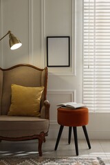 Pouf with books, armchair, lamp and empty frame on white wall in living room. Interior design