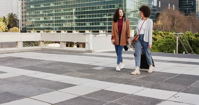 Diverse women friends crossing plaza chatting then waving goodbye one following pulling suitcase