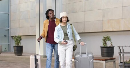 Diverse couple exiting plaza pulling luggage, woman scrolling smartphone, man glancing companion