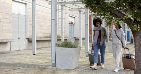 Diverse female friends walking into courtyard from right pulling suitcases to entrance, copy space - Powered by Adobe