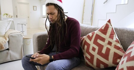 Reading texts African American man in Santa hat sitting on sofa scrolling phone, smiling, laughing - Powered by Adobe