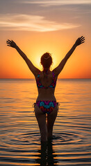 Woman’s back in patterned bikini, arms raised as she walks toward ocean, sun setting behind her