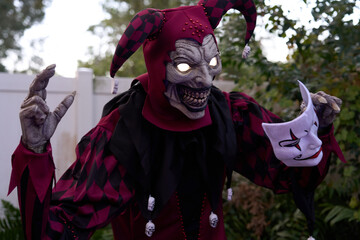 A creepy Halloween jester clown with glowing eyes shows sharp teeth and clawed hands. The red and black harlequin costume holds a white mask in an outdoor yard scene.