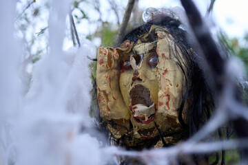 A mutilated doll face with cracked latex and smeared blood hangs in sticky webs. Spooky Halloween prop framed by branches in an outdoor yard.