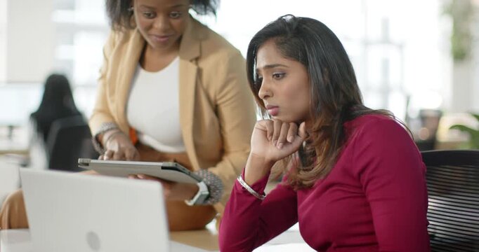 Diverse female coworkers pointing at laptop, guiding data entry and tapping tablet at office desk
