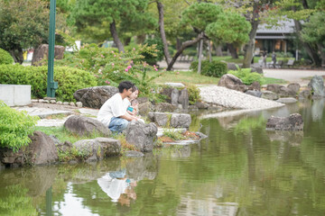 A man and a child are sitting on a rock by a pond. The man is holding a fishing rod and the child is holding a fishing pole. The scene is peaceful and relaxing