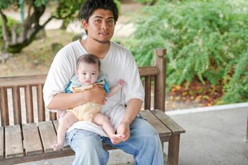 Fototapeta premium A man is holding a baby on a bench. The baby is wearing a yellow outfit. The man is smiling and seems to be enjoying the moment