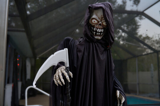 A hooded grim reaper holds a white scythe and shows a toothy grin. Black robe, glowing red eyes, and skeletal hands make a scary outdoor Halloween portrait.