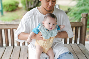 Fototapeta premium A man is holding a baby on a bench. The baby is wearing a blue shirt and a yellow outfit. The scene is peaceful and calm, with the man