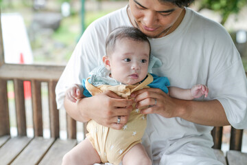 Fototapeta premium A man is holding a baby on a bench. The baby is wearing a yellow outfit and has a blue shirt on. The man is holding the baby's hand and he is taking care of the baby