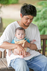 Fototapeta premium A man is holding a baby on a bench. The baby is wearing a blue shirt and a yellow shirt. Scene is warm and loving