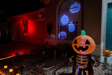 Front yard with a smiling inflatable pumpkin, blue jack-o-lantern window projections, and a red-lit reaper. Festive Halloween home display at night with spooky lights.