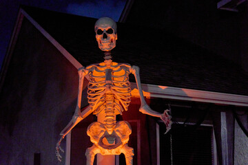 A towering skeleton figure stands by a house porch under orange spotlight. Spooky Halloween yard...