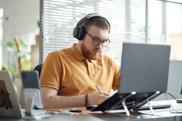 Caucasian young adult man wearing headphones working at desk using laptop and writing in notebook, focused expression, sitting in modern office environment with digital devices visible