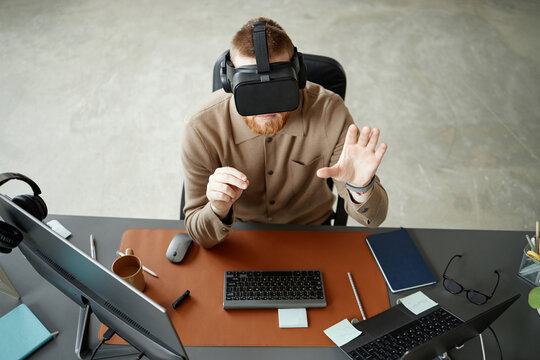 Caucasian young adult man using virtual reality headset while sitting at desk, interacting with digital interface, reaching out with hand, surrounded by computer monitors and office supplies