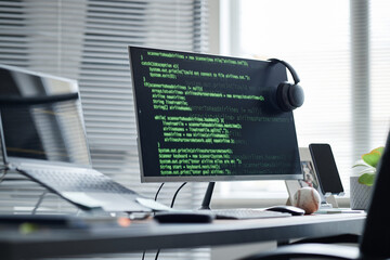 Modern office workspace featuring desktop monitor displaying programming code, wireless headphones resting on screen, laptop and smartphone on desk, baseball and pen holder nearby