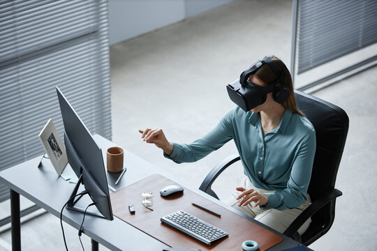 Caucasian young adult woman sitting at desk using virtual reality headset and interacting with digital interface in modern office environment, computer monitor on table