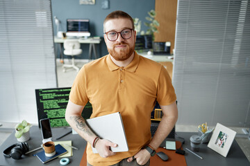 Naklejka premium Portrait of young adult Caucasian man with beard and glasses standing in modern office holding digital tablet, looking confidently at camera with computer monitors in background