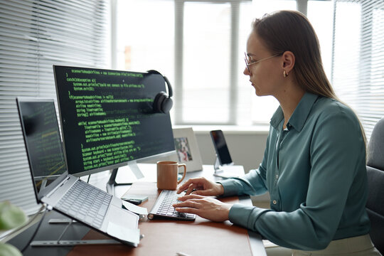 Caucasian young adult woman working at desk using laptop and external monitor displaying programming code, wearing eyeglasses, typing on keyboard in modern office setting