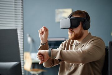 Caucasian young adult man wearing virtual reality headset interacting with digital interface in modern office environment, raising hand and gesturing while sitting at desk