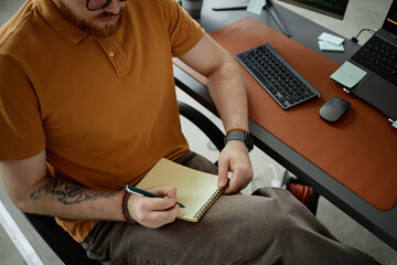 Caucasian young adult man sitting at desk writing in notebook with pen, visible tattoo on forearm, using laptop and keyboard, working in modern office environment