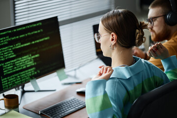 Caucasian young adult woman sitting at desk working on computer with code on screen, stretching arms while male colleague with beard and headphones working in background in modern office
